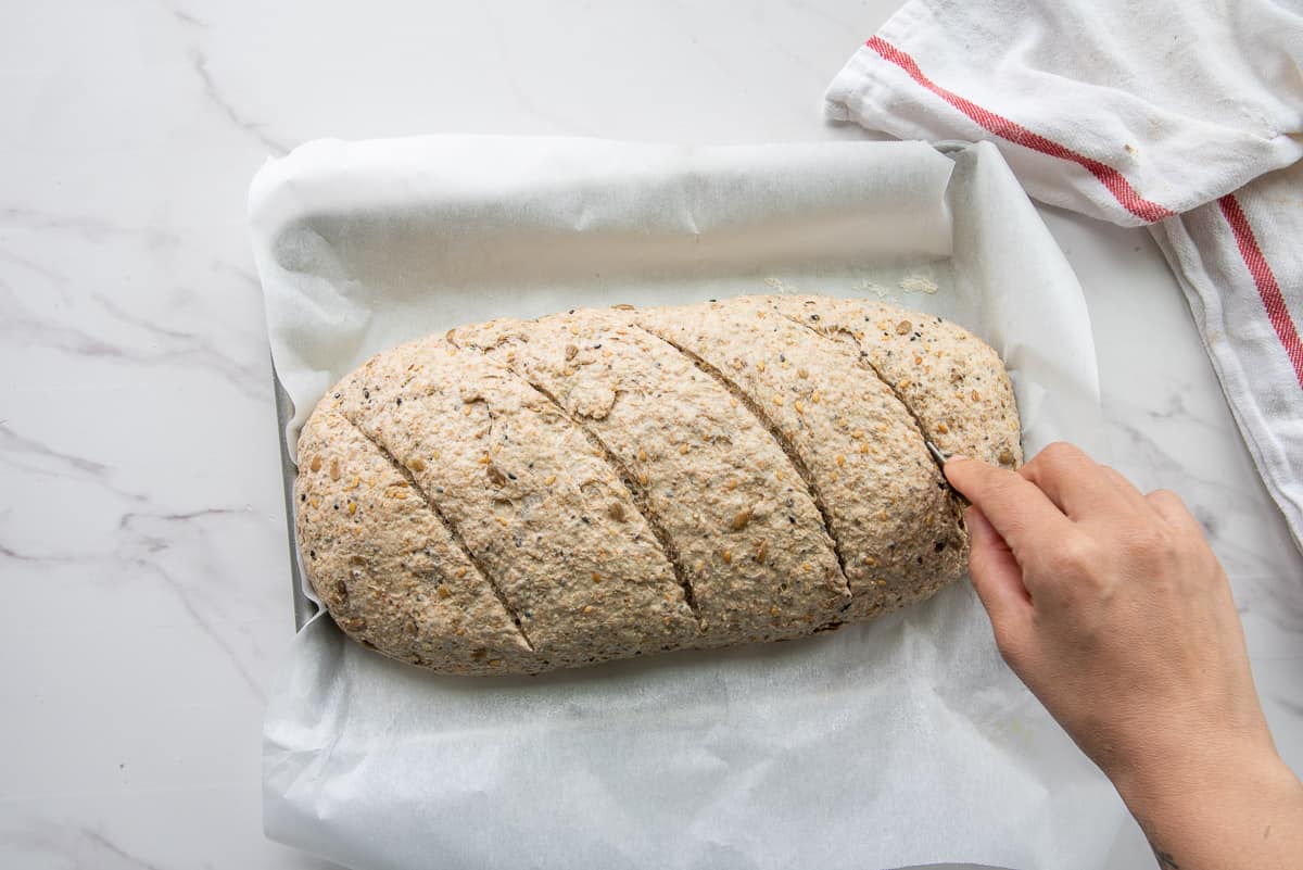A hand uses a razor blade to cut the loaf's top to prevent bursting.