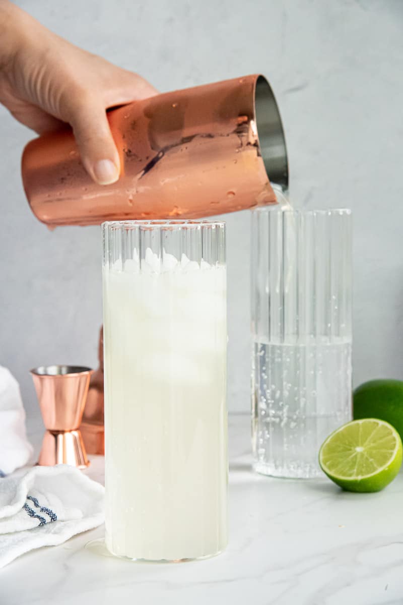 A hand "dirty pours" the drink mixture into a highball glass with club soda.