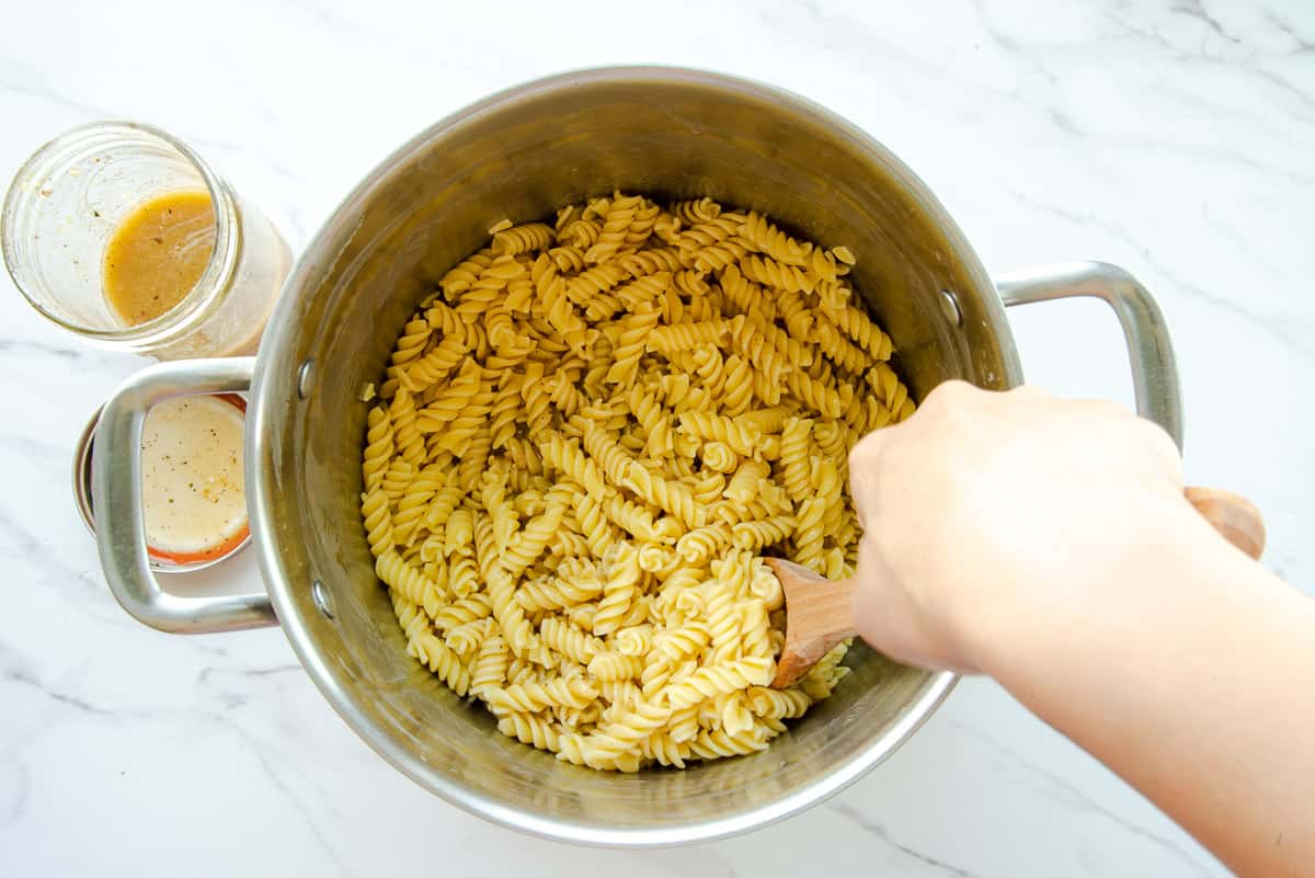 A hand uses a wooden spoon to stir the red wine vinaigrette into a pot of cooked pasta.