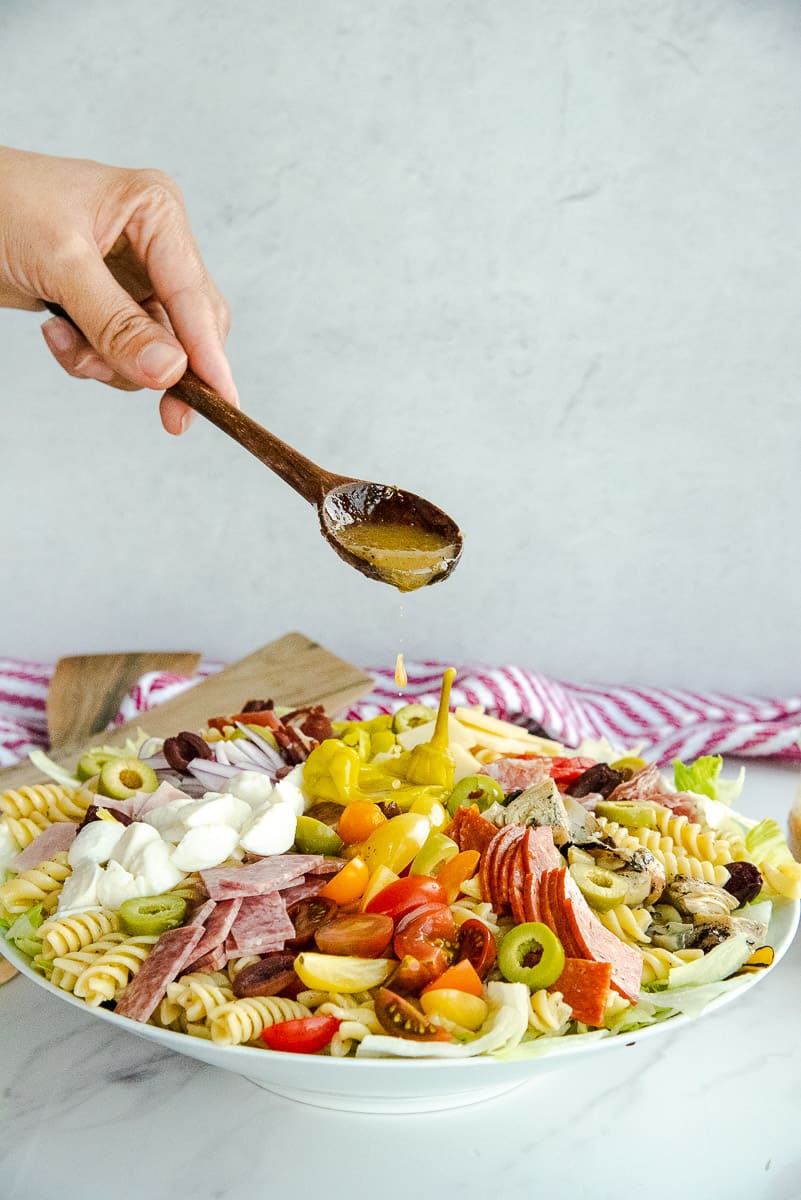 A hand uses a wooden spoon to drizzle red wine vinaigrette over the antipasto pasta salad.