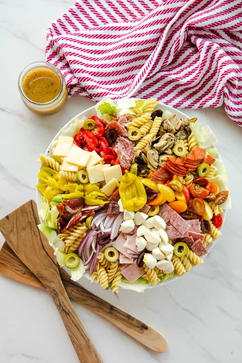 The finished antipasto pasta salad next to salad spoons and a jar of red wine vinaigrette.