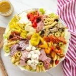 The antipasto pasta salad next to a red and white striped towel a clear glass jar of Italian vinaigrette and two salad spoons.