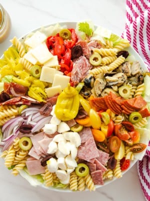 The antipasto pasta salad next to a red and white striped towel a clear glass jar of Italian vinaigrette and two salad spoons.