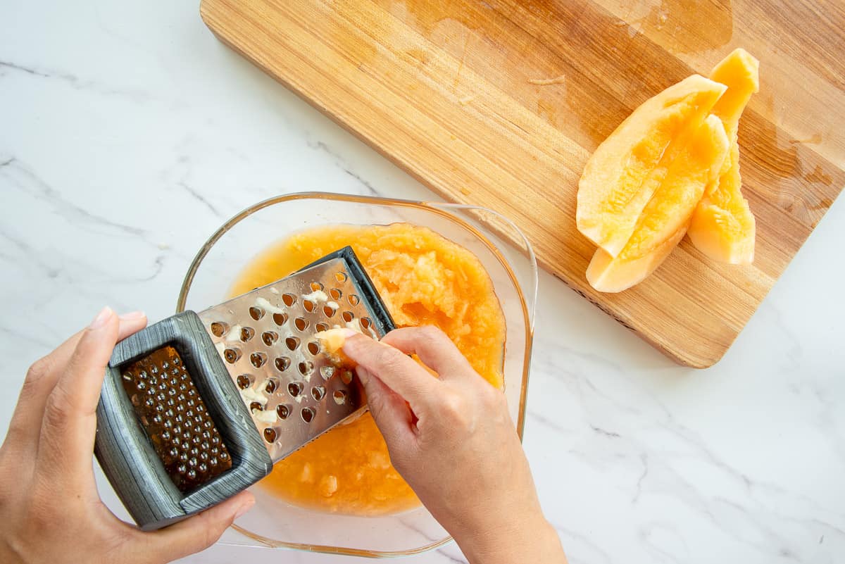 A hand grating cantaloupe slices into a clear glass dish.