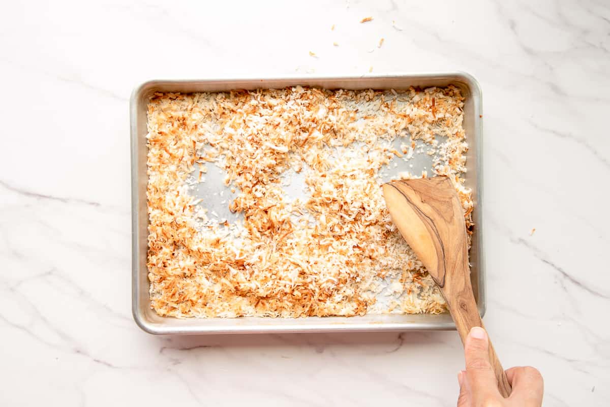 A flat wooden spoon is used to toss the coconut flakes as they are toasted on a silver sheet pan.