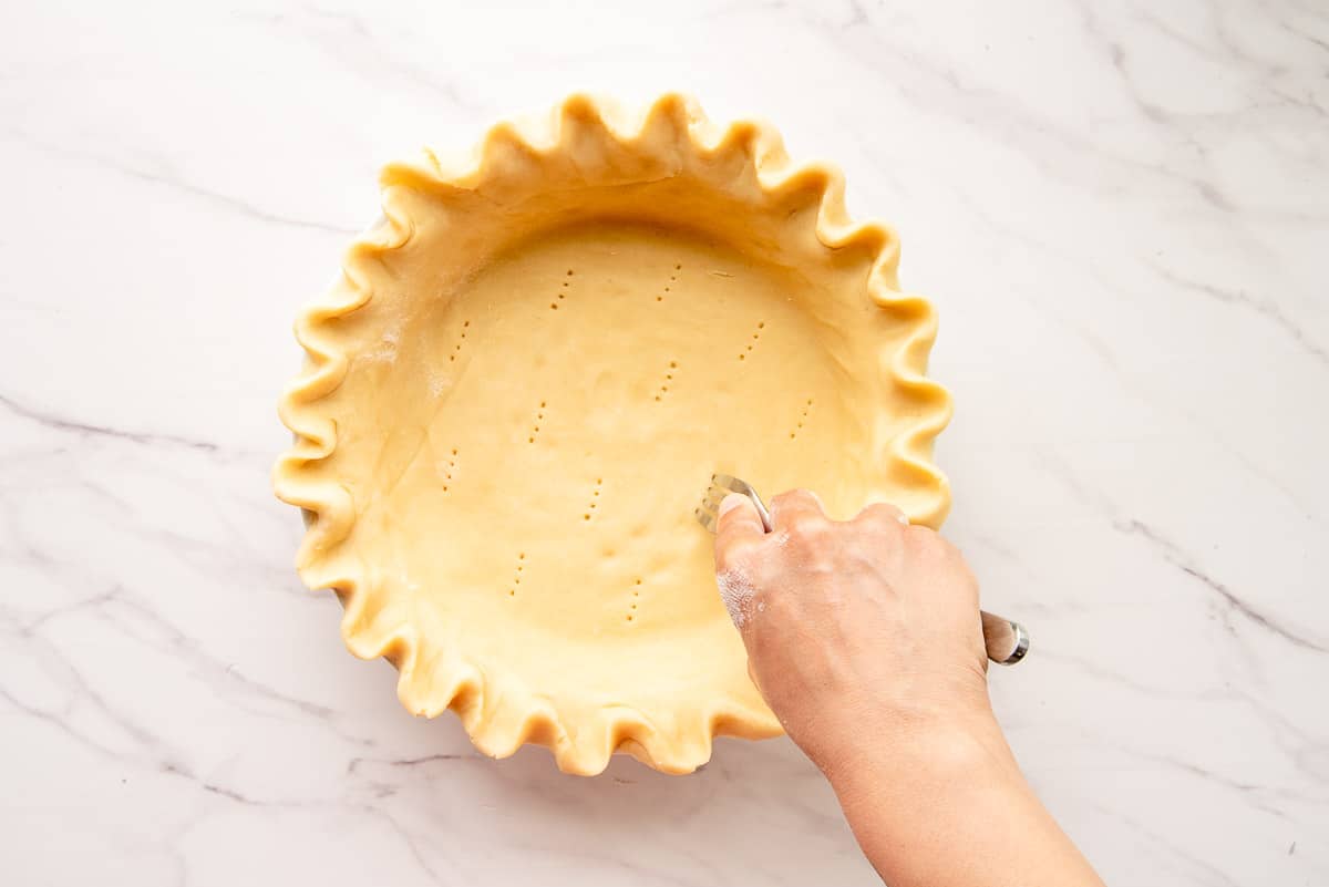 A hand uses a silver fork to pierce holes into the pie dough before blind-baking.