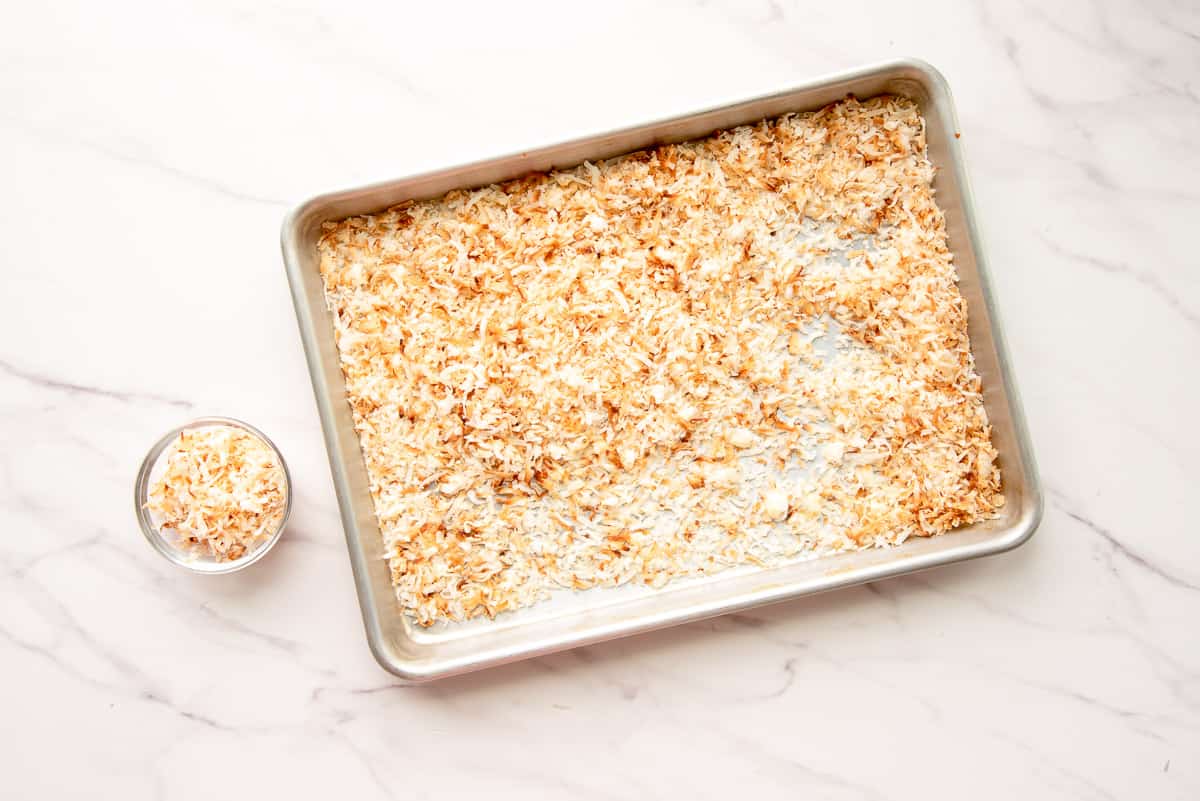 A quarter cup of toasted coconut flakes is removed from the rest of the coconut flakes on a silver sheet tray.