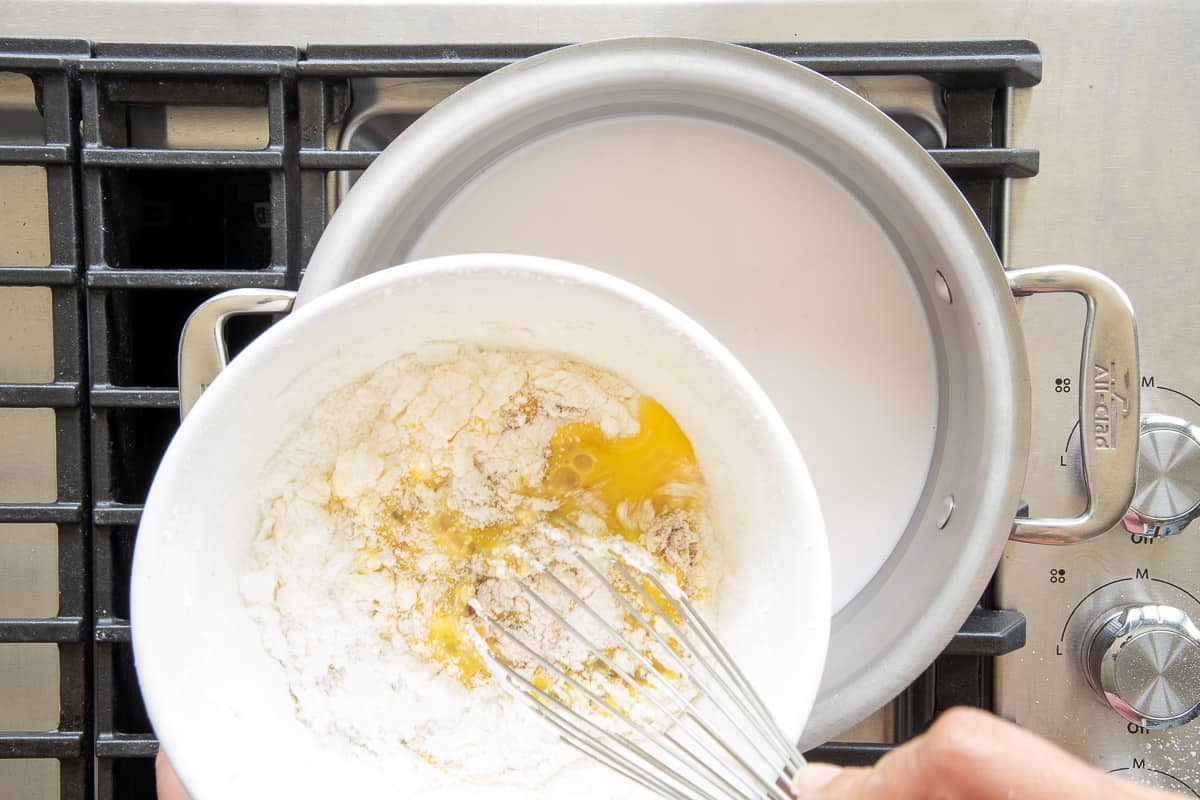 The cornstarch and egg mixture are whisked together in a bowl over a pot of coconut milk on the stove.