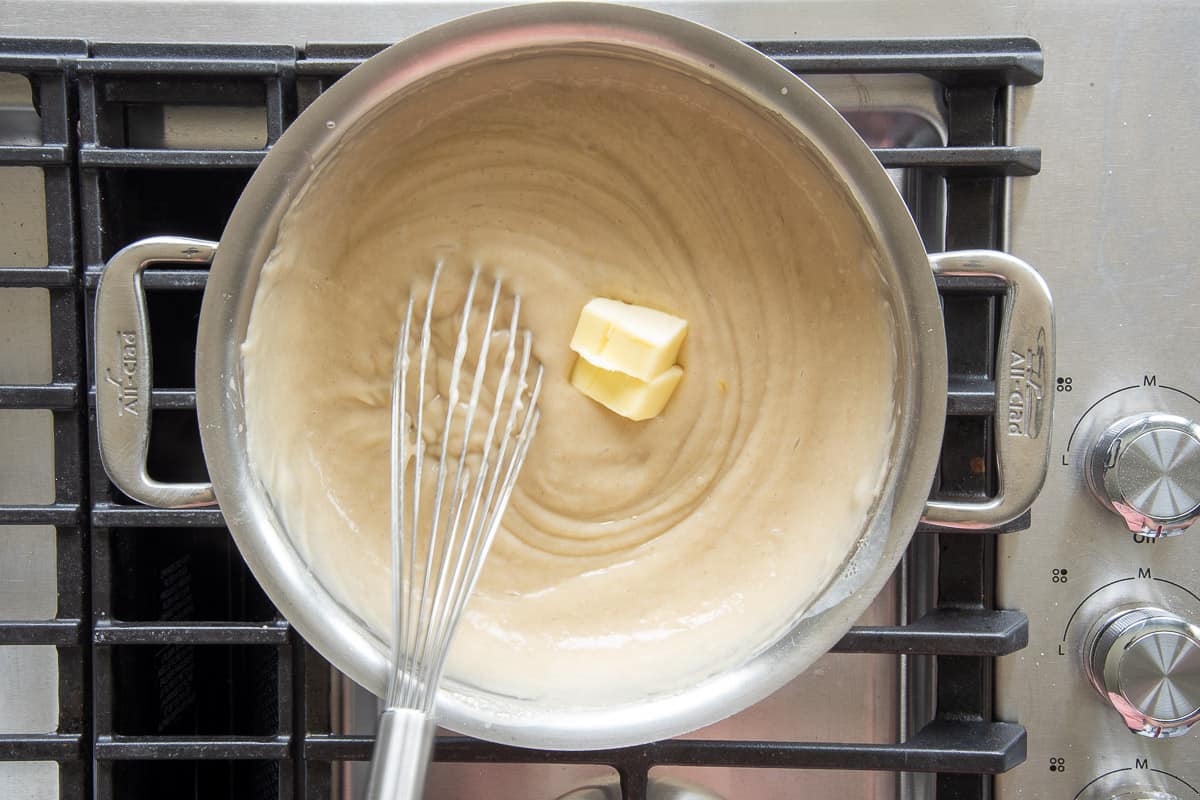 Two slices of unsalted butter are added to the coconut custard in a silver pot with a whisk.