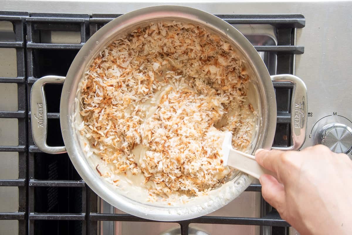 A rubber spatula is used to fold in toasted coconut to the coconut custard in a silver pot on a stove.