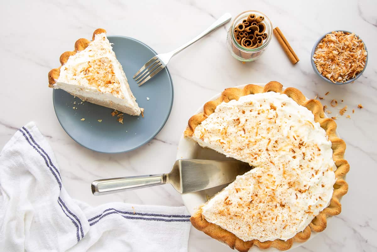 The finished Coconut Rum Cream Pie garnished with toasted coconut flakes, cinnamon, and two cinnamon sticks the Coquito cream pie with the sliced remove to the left next to a jar of cinnamon sticks and a blue bowl of toasted coconut flakes.