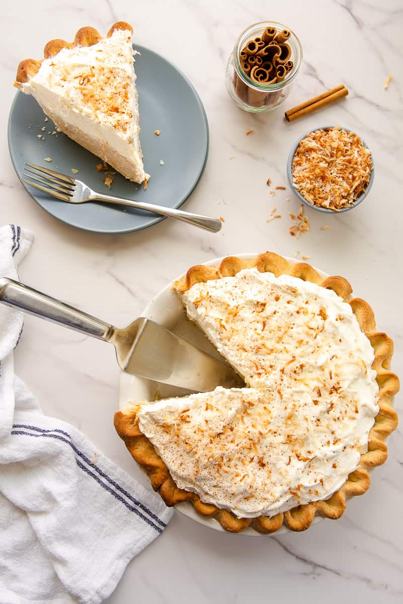 The Coconut Rum Cream Pie next to a blue plate with a slice of pie on it, a jar of cinnamon sticks, and a bowl of toasted coconut flakes.
