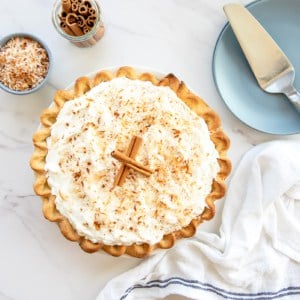 The finish coquito cream pie garnished with two cinnamon sticks next to a bowl of toasted coconut, a jar of cinnamon sticks and two blue plates with a silver cake server on top.