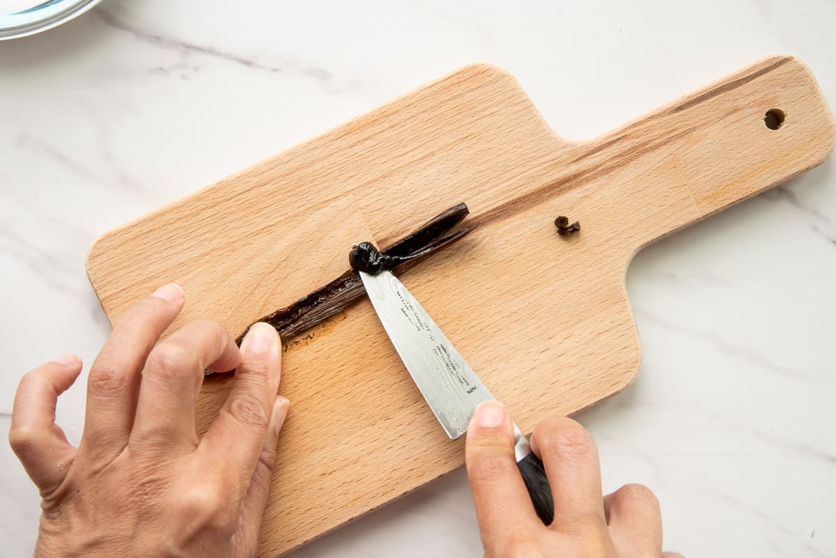 Hands hold a vanilla bean pod split open while the other hand scrapes the vanilla bean paste from the pod on a wooden cutting board.