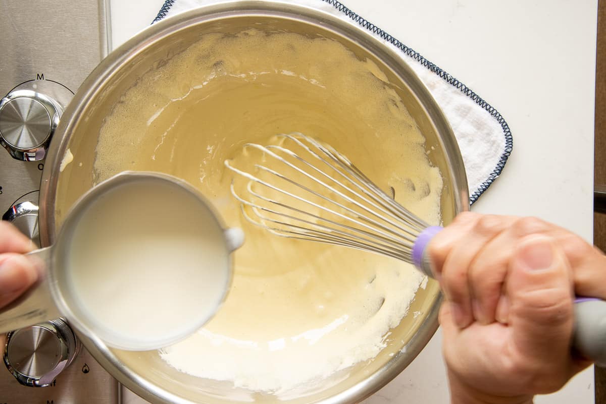 The hot milk mixture is added to the egg and sugar mixture with a whisk in a metal mixing bowl.
