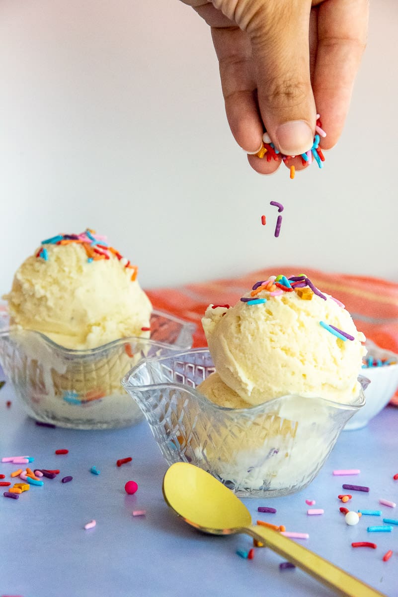 A hand sprinkles rainbow jimmies on two scoops of double vanilla frozen custard in glass dessert bowls.