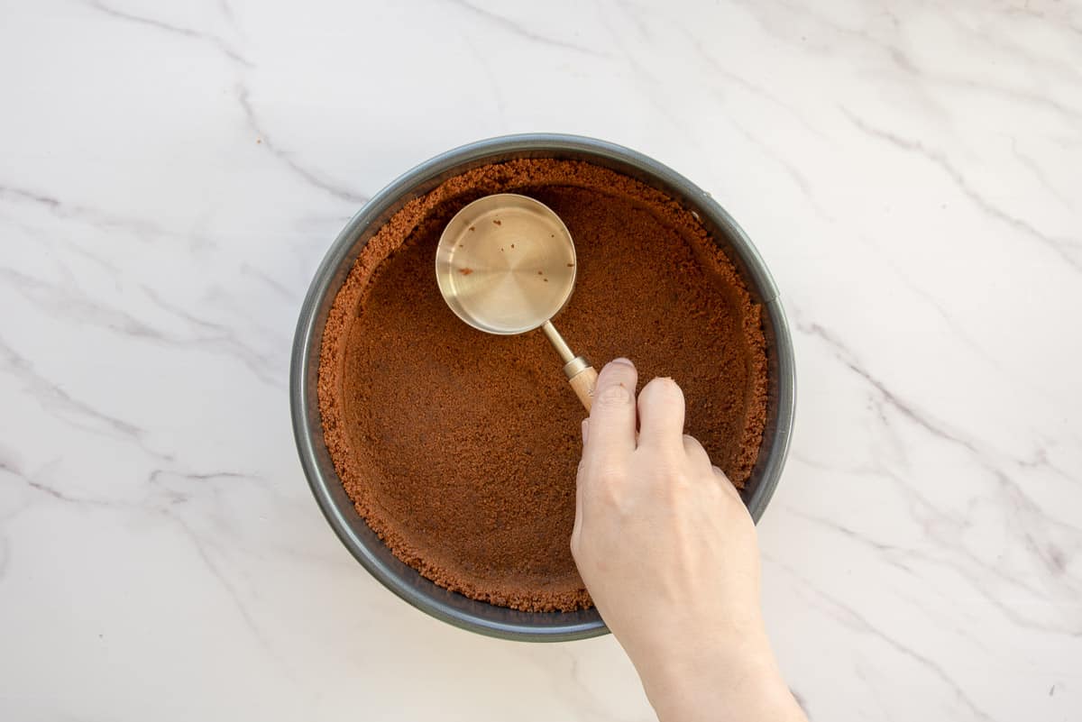 A hand uses a measuring cup to form the gingersnap crust.