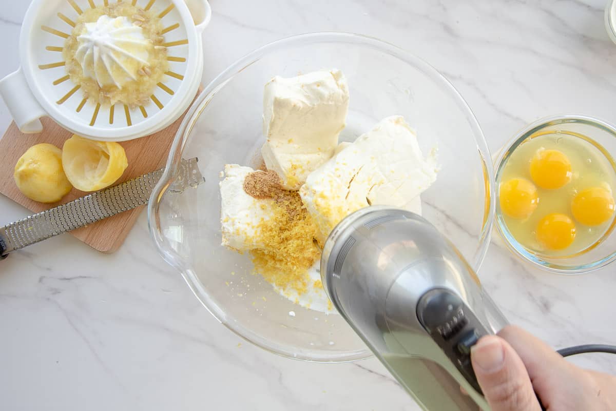 An electric mixer is used to blend the ingredients for the lemon meringue cheesecake batter in a clear glass bowl.