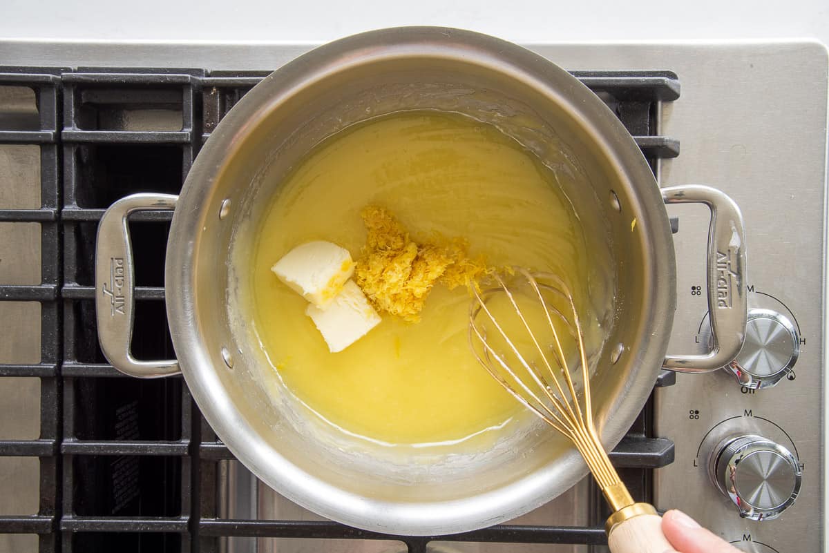 Butter and lemon zest are added to the lemon curd mixture in a silver pot on the stove.