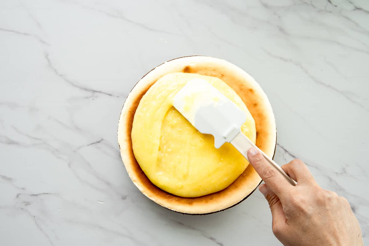A hand uses a rubber spatula to spread the lemon curd mixture on top of the bake cheesecake