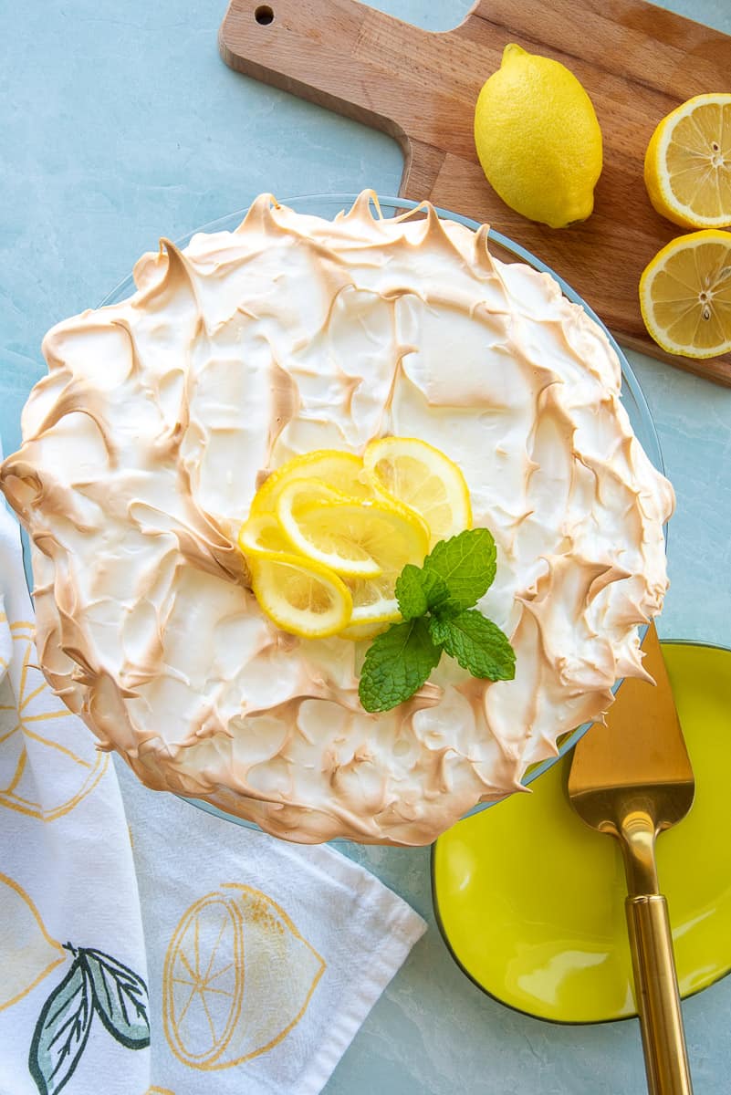 A lemon meringue cheesecake garnished with lemon slices fresh mint next to a wooden cutting board with one whole lemon and one lemon cut in half.