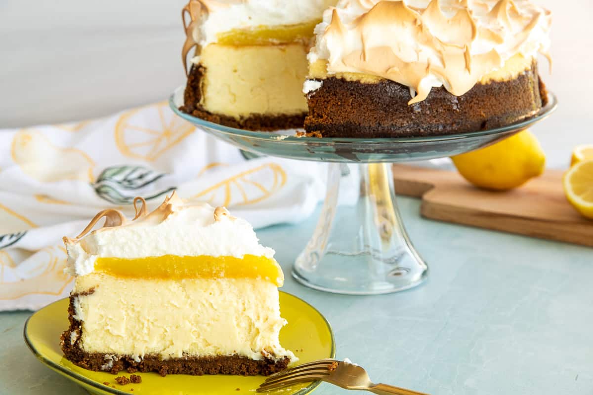 A slice of lemon meringue cheesecake on a white on a yellow plate in front of a clear cake stand with the rest of the cheesecake on it.
