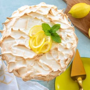 The lemon meringue cheesecake garnished with lemon slices fresh mint on a cake stand next to a cutting board with cut lemons on it.