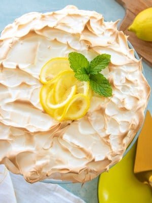 The lemon meringue cheesecake garnished with lemon slices fresh mint on a cake stand next to a cutting board with cut lemons on it.