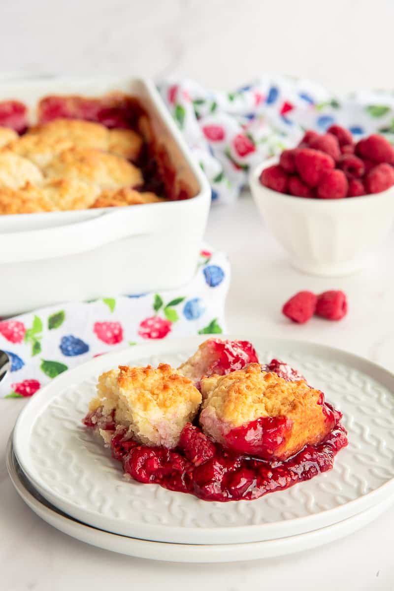 A serving of raspberry cobbler on a white plate in front of the baking dish of cobbler and a bowl of fresh raspberries.