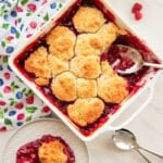 Square white ceramic baking dish with raspberry cobbler. A serving is removed and on a white plate at bottom left of image.