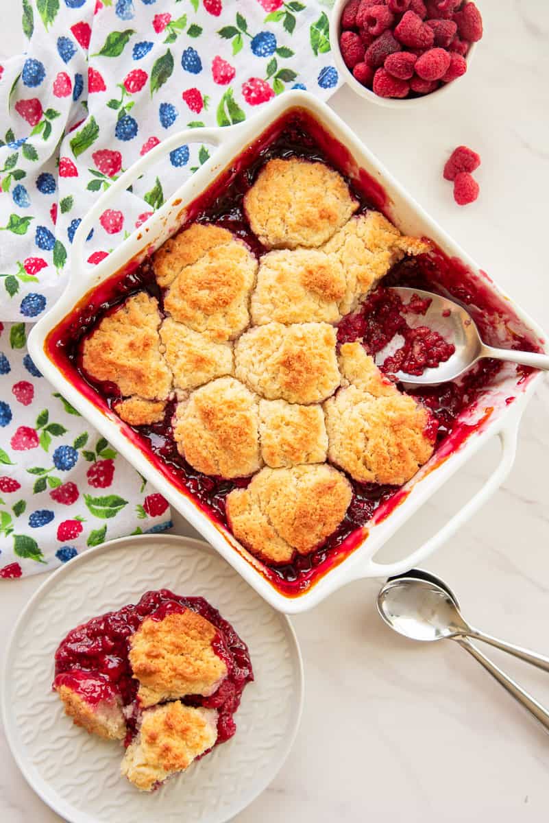 Square white ceramic baking dish with raspberry cobbler. A serving is removed and on a white plate at bottom left of image.