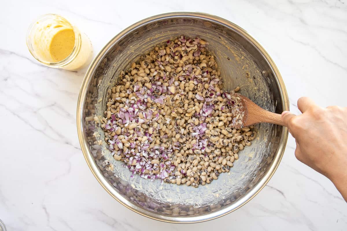 A hand stirs the ginger-garlic vinaigrette into the black eyed peas along with red onions.