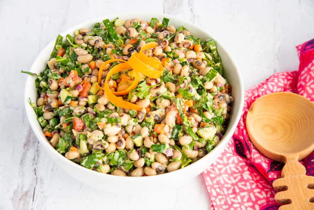 White bowl filled with black eyed peas salad with collard greens garnished with ribbons of carrots next to a wooden spoon.