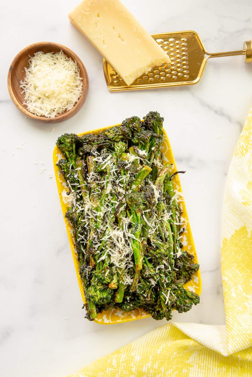 Charred spicy broccolini on a yellow and white decorative platter next to a wooden bowl filled with Parmesan cheese and a block of Parmesan cheese on top of a gold grater.