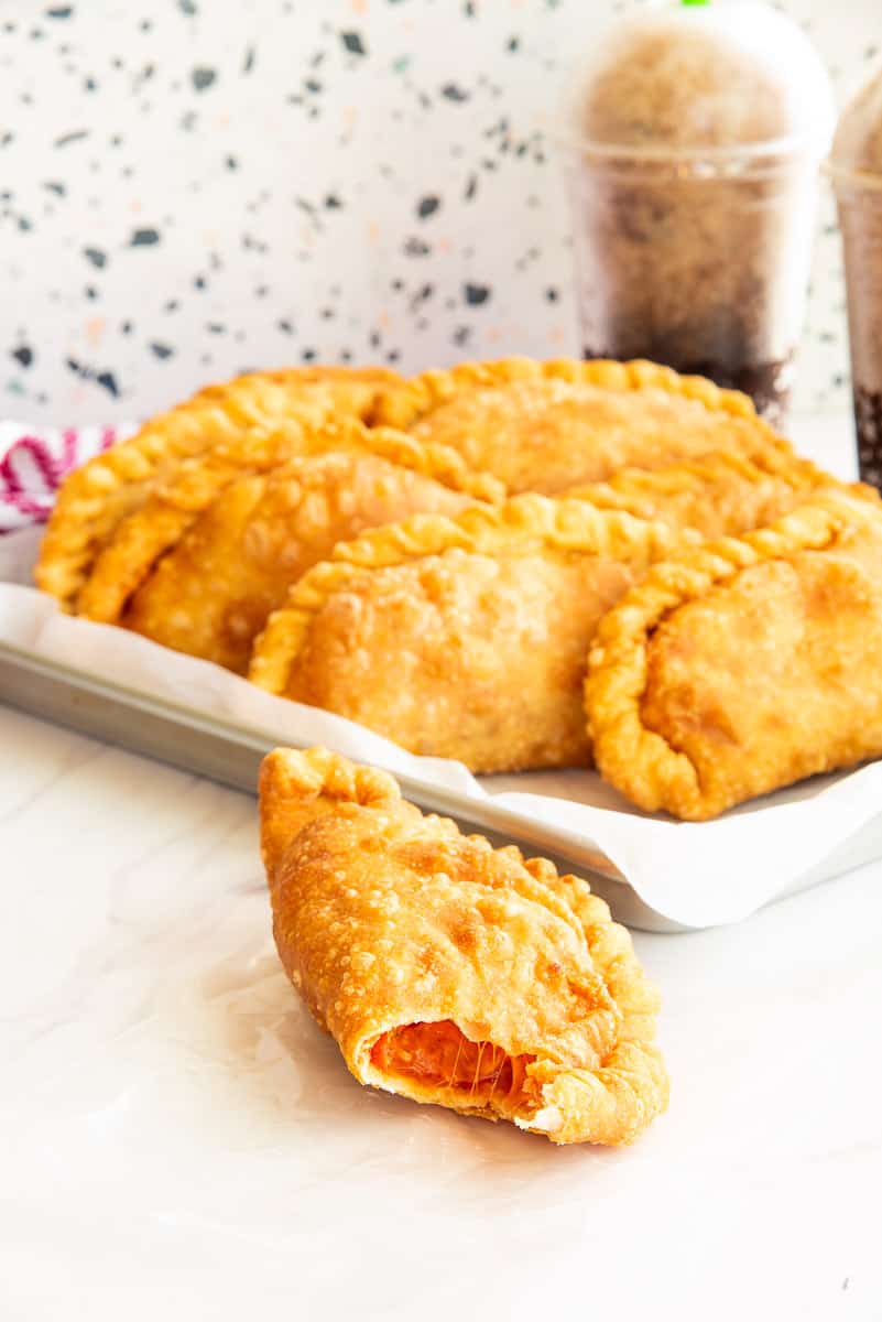 A pizza empanada with a bite removed is on a piece of wax paper in front of a sheet pan with the rest of the empanadas on it.