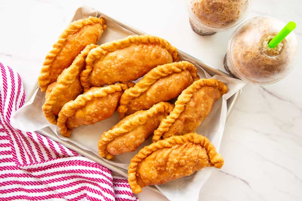 The fried pizza empanadas are on a parchment-lined sheet pan next to two cola slushies.