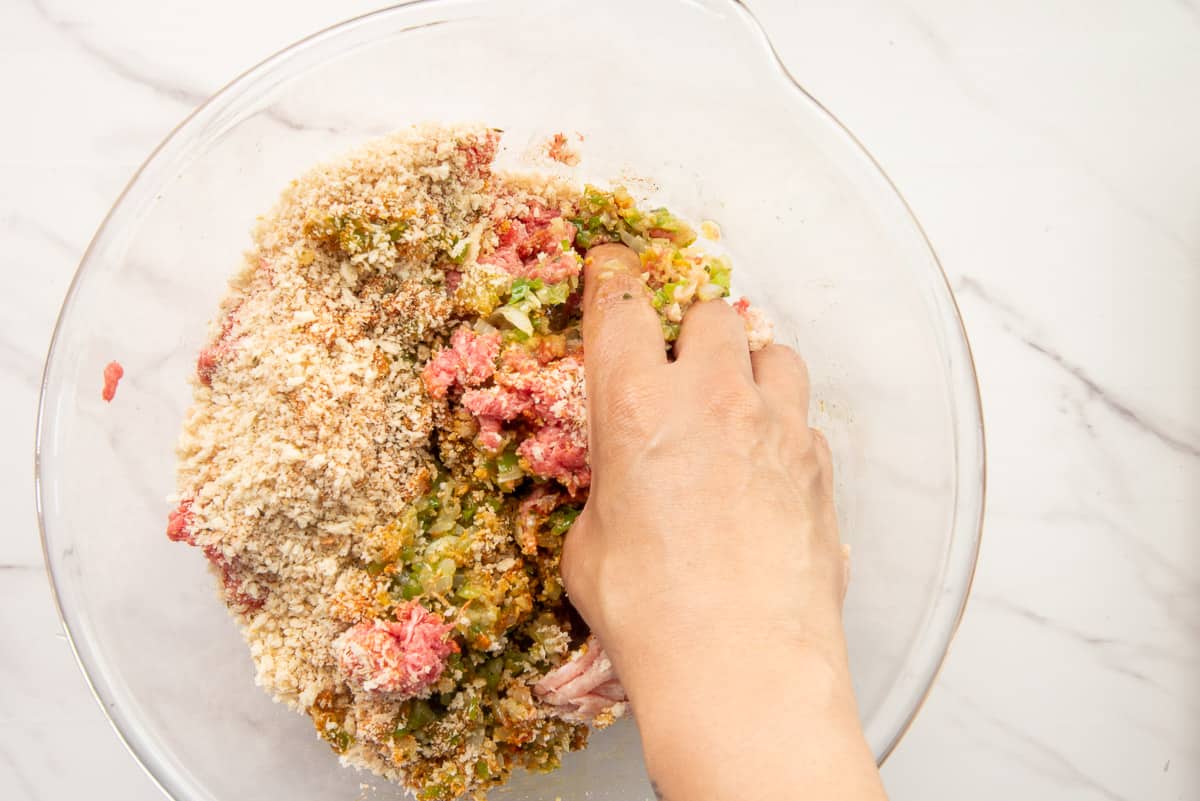 A hand mixes the meatball ingredients together in a clear glass bowl.
