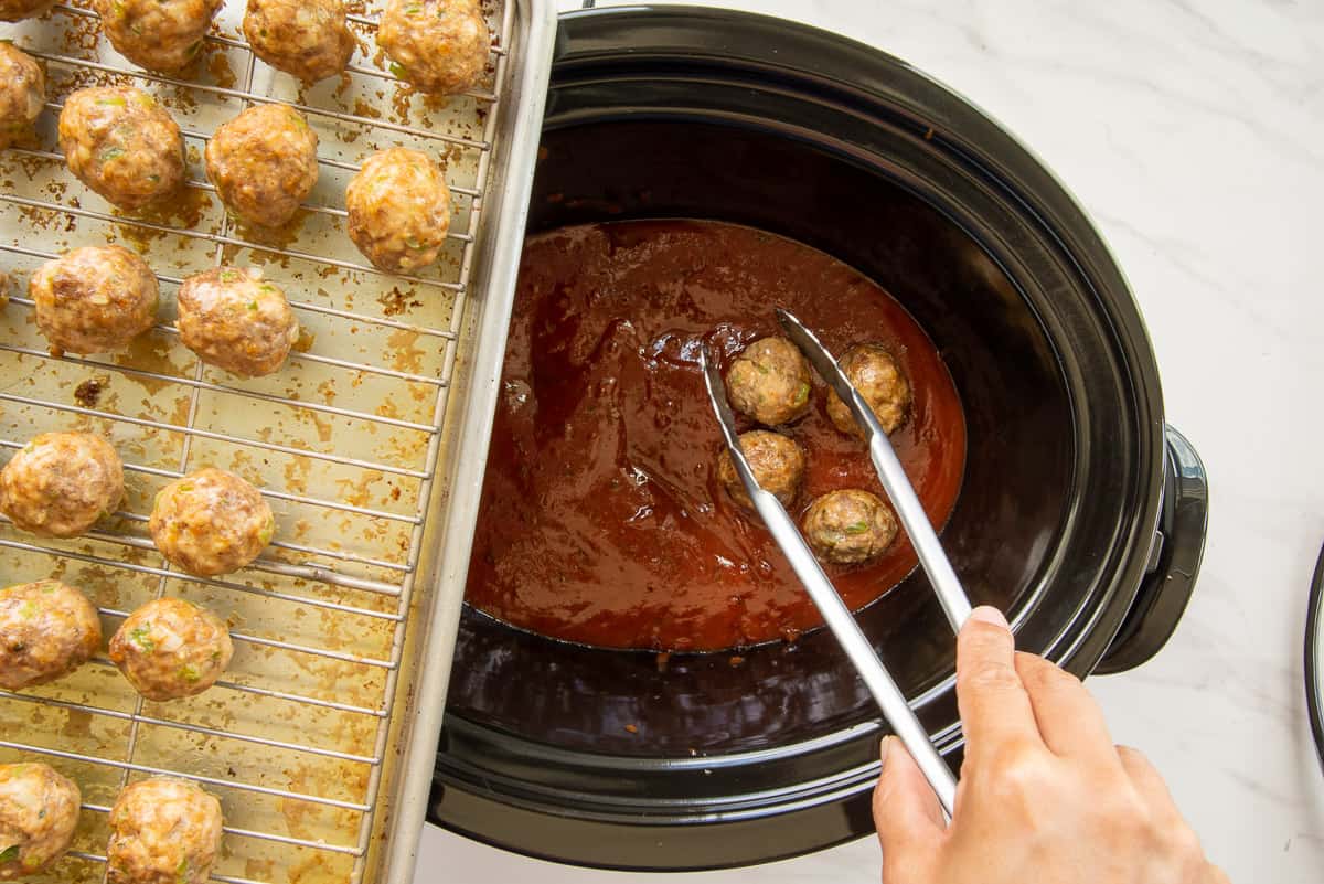 The baked meatballs are added to the sauce in a slow cooker using tongs.