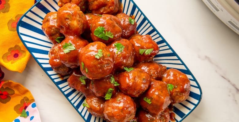 Party meatballs in guava sauce on a blue and white platter next to a white slow cooker and a yellow plate with meatballs on it.