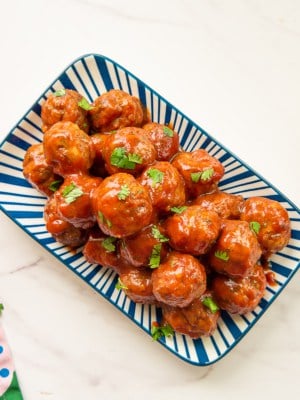 Party meatballs in guava sauce on a blue and white platter next to a colorful towel.