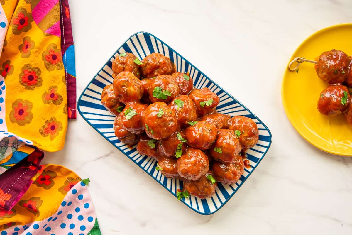 Party meatballs in guava sauce on a blue and white platter next to a colorful towel.