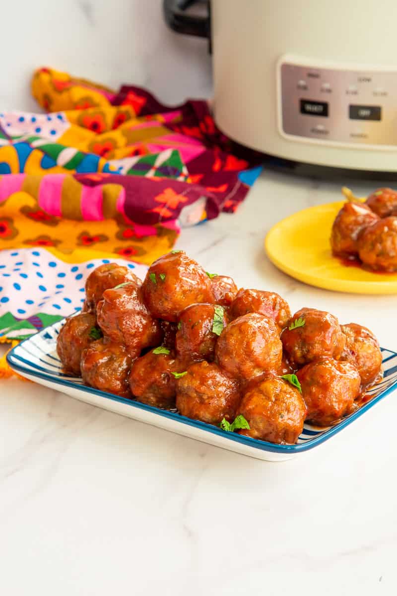 A stack of party meatballs in guava sauce on a blue and white platter in front of a white slow cooker.