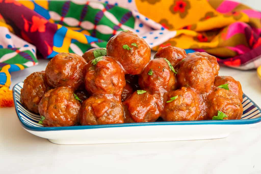 Party meatballs in guava sauce on a white platter in front of a colorful kitchen towel.