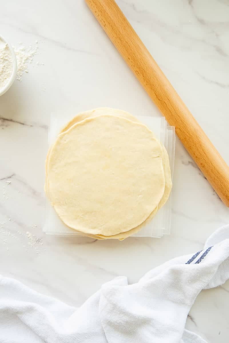 Empanada dough stacked next to a wooden rolling pin and a blue and white kitchen towel