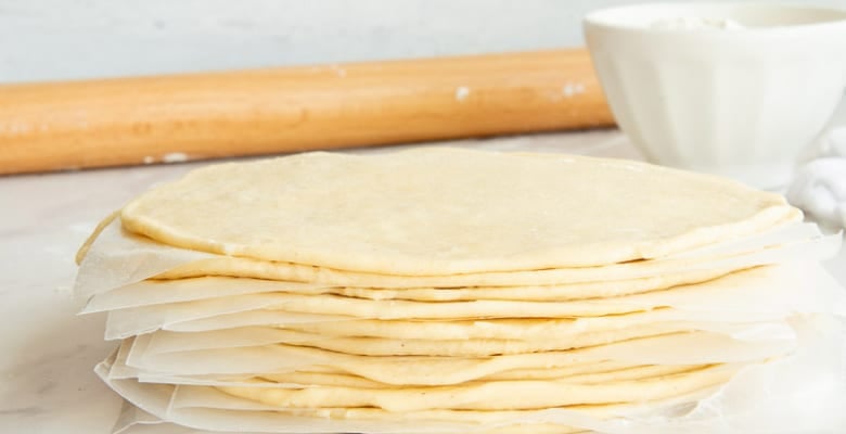 Empanada dough discs stacked in front of a wooden rolling pin and white bowl.