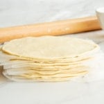 The discs are stacked in front of a white bowl of flour and a wooden rolling pin.