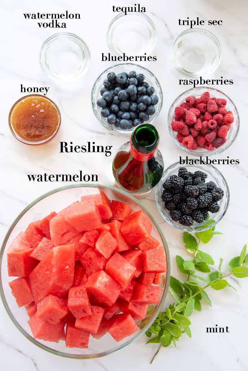 Ingredients needed to prepare watermelon sangria on a white countertop are labeled with black text.