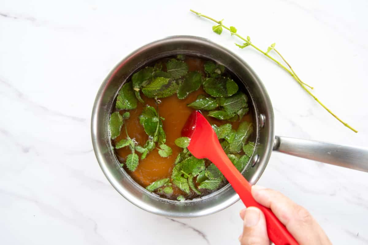 The ingredients for the honey-mint simple syrup are combined in a silver pot with a red rubber spatula.