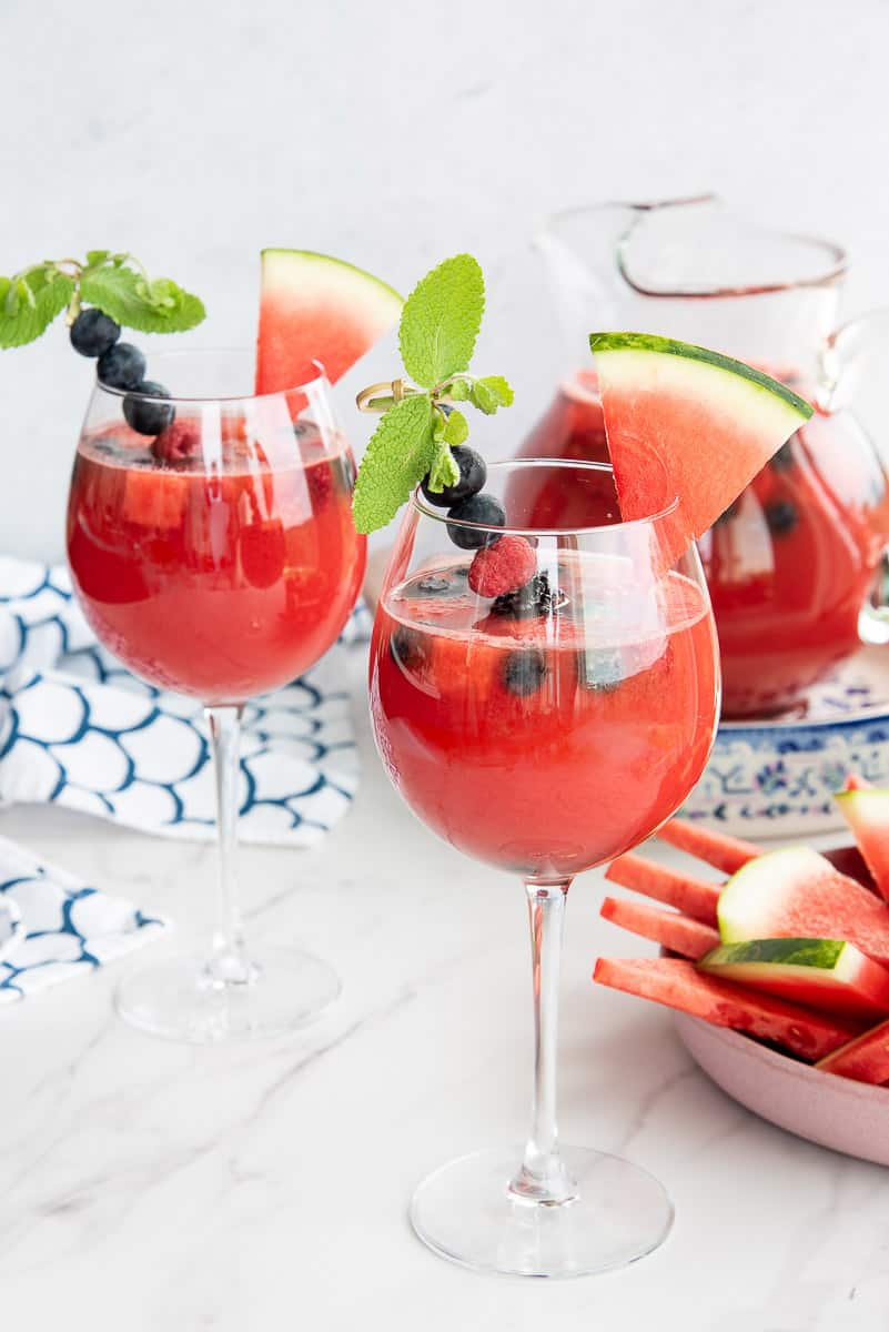 Wine glasses filled with watermelon sangria are garnished with a sprig of mint, fresh berries, and a triangle of watermelon.