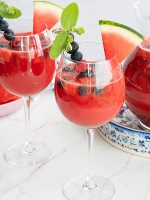 Two wine glasses filled with watermelon sangria garnished with a skewer of berries mint and a triangle of watermelon in front of a pitcher with the rest of the watermelon sangria.